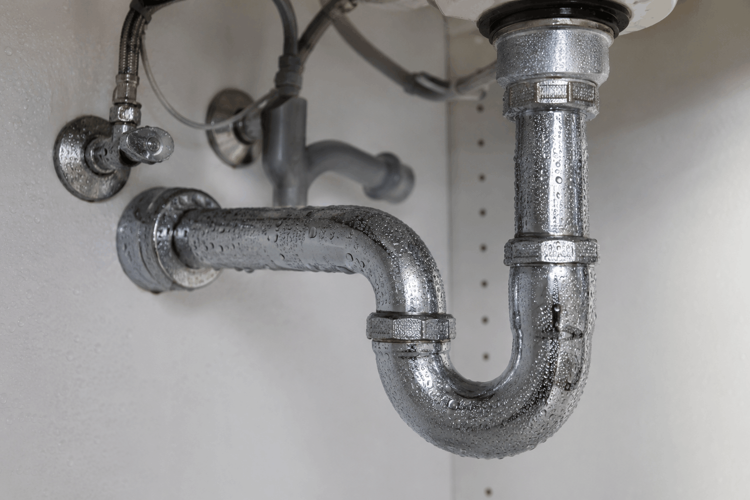 Water droplets covering plumbing pipes beneath a sink cabinet.